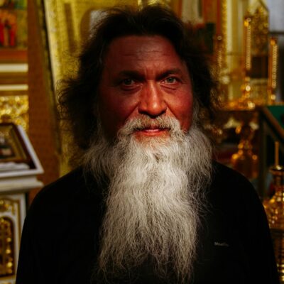 Peaceful senior Orthodox priest with gray beard in cassock standing in church near golden icons and frescoes and looking at camera